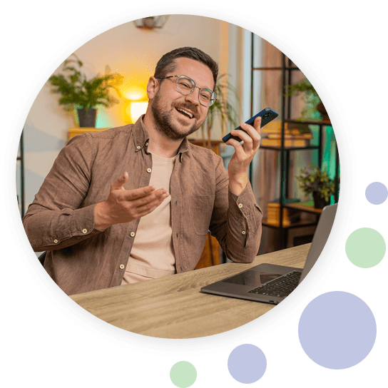 Man zit lachend aan tafel met laptop en voert een gesprek via zijn smartphone.